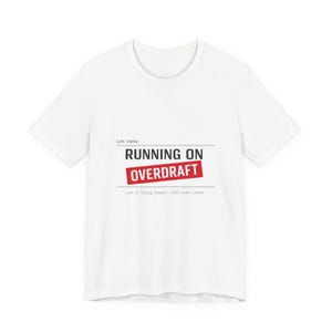 Könnte beinhalten: Weißes T-Shirt mit dem Text "RUNNING ON OVERDRAFT" in Schwarz und Rot. Das Shirt enthält auch die Sätze "Life status:" und "cost of living request: still under review" in Schwarz. Ein lässiges T-Shirt für den Alltag.