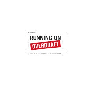 Könnte beinhalten: Ein weißer Aufkleber mit dem Text "RUNNING ON OVERDRAFT" in Schwarz und Rot. Die Wörter sind gestapelt, mit "OVERDRAFT" in einem roten Rechteck. Darunter steht in kleinerem Text "cost of living request: still under review."