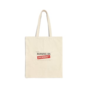 Könnte beinhalten: Cremefarbene Tragetasche mit langen Henkeln. Die Tasche zeigt den Text "RUNNING ON OVERDRAFT" in einem roten Rechteck, mit kleinerem Text darüber und darunter. Die Tasche ist aus robustem Stoff gefertigt.