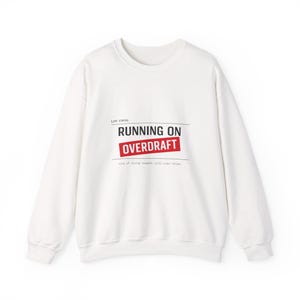 Könnte beinhalten: Weißes Sweatshirt mit Rundhalsausschnitt und dem Text "RUNNING ON OVERDRAFT" in Schwarz und Rot. Darunter steht "cost of living request: still under review" in Schwarz. Das Sweatshirt hat lange Ärmel.