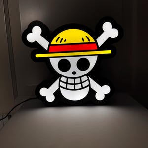 Può includere: Un'insegna luminosa di One Piece Jolly Roger nera, bianca, gialla e rossa. Il design presenta un teschio con un cappello di paglia e ossa incrociate. L'insegna è illuminata e si trova su una superficie bianca.