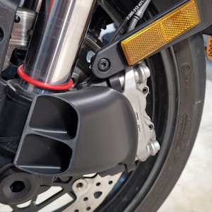 Può includere: Primo piano del gruppo ruota anteriore di una moto. L'immagine evidenzia la forcella anteriore argentata, il parafango nero e i componenti dei freni. È visibile un catarifrangente di sicurezza arancione riflettente. Lo pneumatico è nero con un disegno del battistrada visibile.
