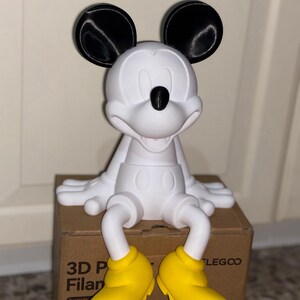 Puede incluir: Figura de Mickey Mouse impresa en 3D, blanca con orejas y nariz negras, y zapatos amarillos. La figura está sentada en una caja de cartón. Es un objeto decorativo.