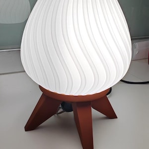 Può includere: Una lampada da tavolo bianca a forma di uovo con un motivo a spirale strutturato. La lampada poggia su una base a treppiede marrone scuro. Il paralume misura circa 30 cm di diametro e 46 cm di altezza.