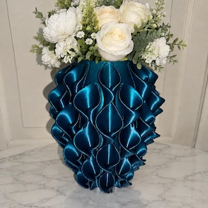Op de afbeelding: Een diep teal-kleurige vaas met een gestructureerd, herhalend bloembladachtig patroon aan de buitenkant. De vaas is gevuld met een arrangement van witte en crèmekleurige kunstmatige rozen, kleine witte vulbloemen en groene bladeren.
