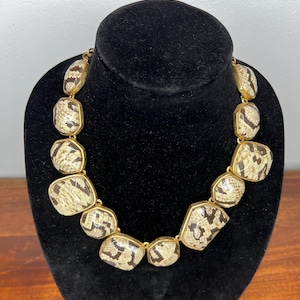 Puede incluir: Un collar llamativo con grandes cuentas geométricas. Las cuentas tienen un patrón de piel de serpiente en beige, marrón y negro, engastadas en metal dorado. Un accesorio audaz.