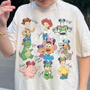 May include: Cream-colored t-shirt featuring a colorful graphic of Toy Story characters, including Woody, Jessie, Buzz Lightyear, and others. Each character is depicted with a Mickey Mouse ear headband. The names of the characters are written in a handwritten font.