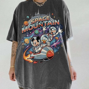 May include: Dark gray t-shirt featuring a colorful graphic of Mickey Mouse, Donald Duck, and Goofy in space suits riding a rocket ship. The words "SPACE MOUNTAIN" are printed above the characters, with planets and stars in the background.
