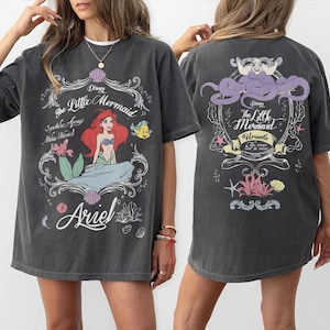 Może przedstawiać: Ciemnoszara koszulka z nadrukiem z przodu przedstawiającym Ariel z Małej Syrenki Disneya, z napisem "The Little Mermaid" i "Ariel". Tył koszulki przedstawia Urszulę oraz napisy "The Little Mermaid" i "Ursula".