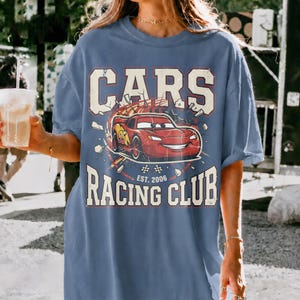 May include: A blue t-shirt featuring the text "CARS RACING CLUB" with a graphic of a red race car. The design includes the text "EST. 2006". The shirt has a relaxed fit and short sleeves.