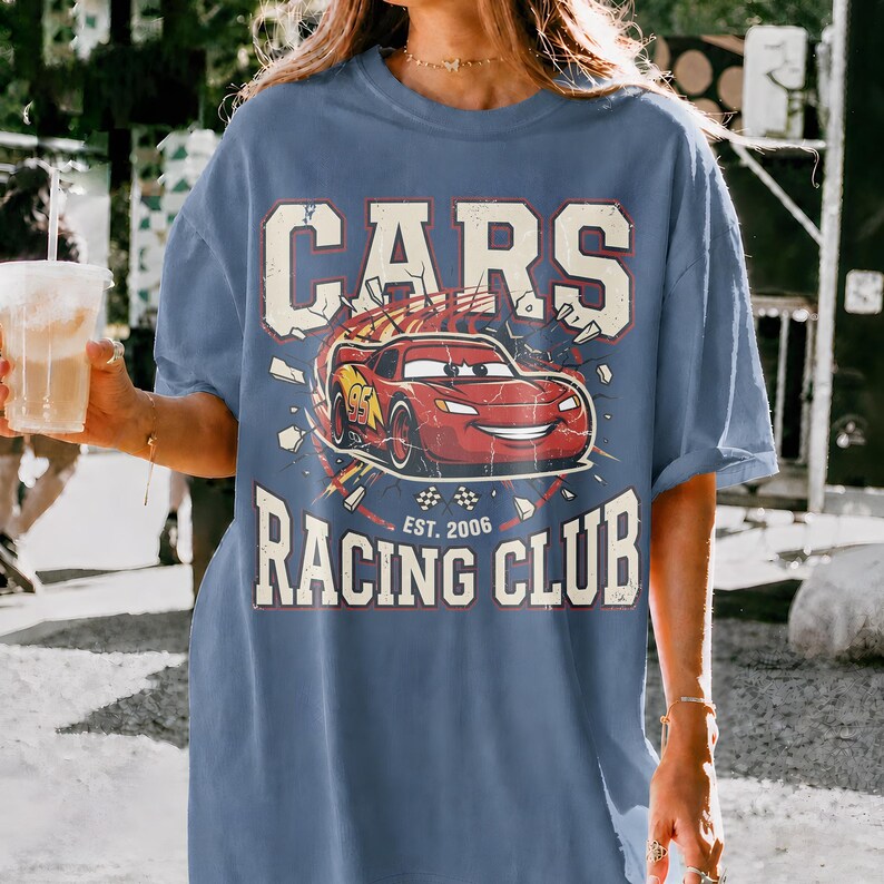 May include: A blue t-shirt featuring the text "CARS RACING CLUB" with a graphic of a red race car. The design includes the text "EST. 2006". The shirt has a relaxed fit and short sleeves.