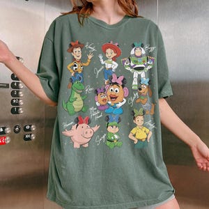 May include: Green t-shirt featuring a colorful graphic of Toy Story characters, including Woody, Jessie, Buzz Lightyear, and others. The design includes character names in a playful font. The shirt has a relaxed fit and short sleeves.