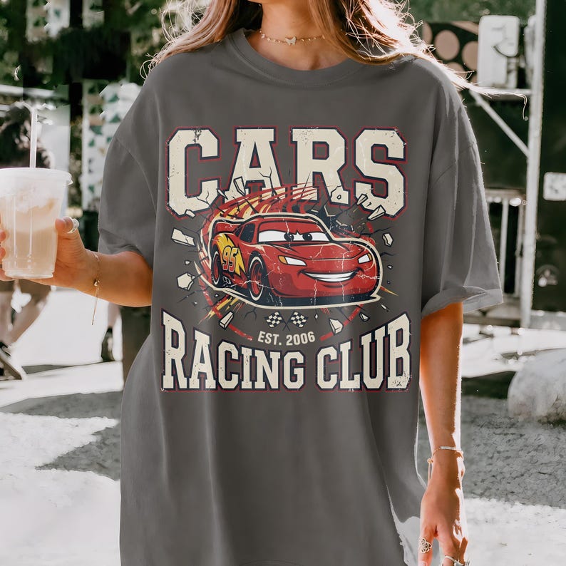 May include: Gray t-shirt featuring a graphic of the animated character Lightning McQueen from the movie Cars. The design includes the text "CARS RACING CLUB EST. 2006" in a vintage style.