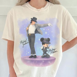 Può includere: T-shirt color crema con una grafica di Michael Jackson e Topolino in abito, in posa. I testi "Michael Jackson" e "Mickey Mouse" sono scritti in corsivo. Lo sfondo ha una combinazione di colori viola e blu con stelle.