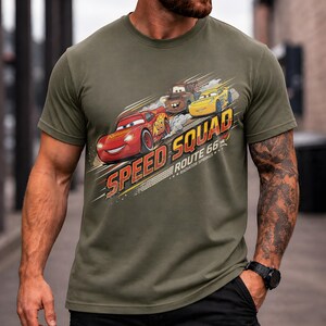 Op de afbeelding: Olijfgroen t-shirt met de tekst "Speed Squad Route 66" en illustraties van Bliksem McQueen, Takel en een gele auto. Het ontwerp heeft een diagonale lay-out met een retro esthetiek.