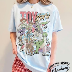 May include: A light blue short-sleeve t-shirt featuring a graphic print of Toy Story characters including Woody, Buzz Lightyear, Jessie, Rex, Slinky Dog, Bullseye, and Hamm. The text reads Toy Story, Jessie Since 1995, and character names.