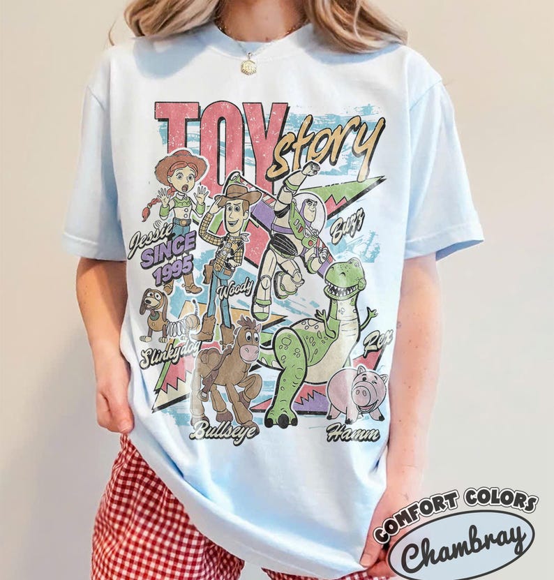 May include: A light blue short-sleeve t-shirt featuring a graphic print of Toy Story characters including Woody, Buzz Lightyear, Jessie, Rex, Slinky Dog, Bullseye, and Hamm. The text reads Toy Story, Jessie Since 1995, and character names.