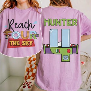 以下が含まれることがあります： 前面に「Reach FOUR The Sky」の文字が入った薄紫色のTシャツ。漫画のカウボーイとカラフルな文字が特徴です。Tシャツの背面には、宇宙船に似た緑と紫のグラフィックの上に「HUNTER」と表示されています。