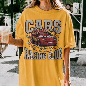 May include: Yellow t-shirt with a Cars Racing Club graphic. The design features the word "CARS" in a gray, distressed font above a red race car illustration. The text "RACING CLUB" is below the car, with "EST. 2006" at the bottom.