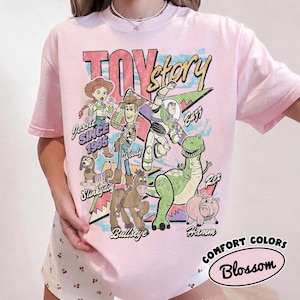 May include: A light pink t-shirt featuring a graphic print of Toy Story characters including Jessie, Woody, Buzz Lightyear, Slinky Dog, Bullseye, Rex, and Hamm. The text reads Toy Story, Since 1995, Jessie, Woody, Buzz, Rex, Slinky Dog, Bullseye, and Hamm.