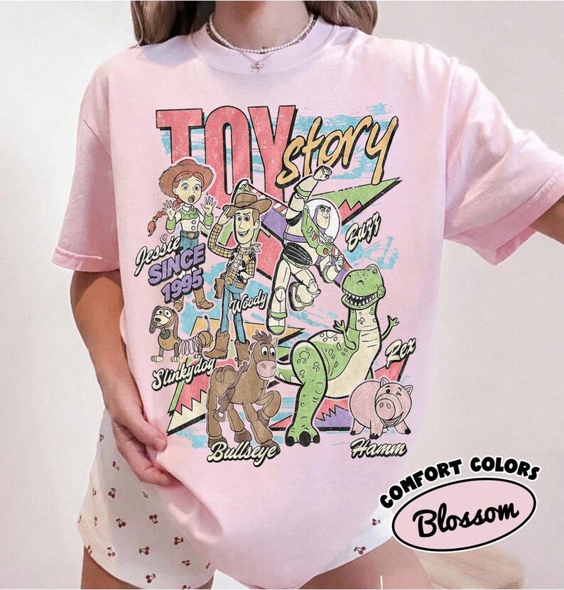 May include: A light pink t-shirt featuring a graphic print of Toy Story characters including Jessie, Woody, Buzz Lightyear, Slinky Dog, Bullseye, Rex, and Hamm. The text reads Toy Story, Since 1995, Jessie, Woody, Buzz, Rex, Slinky Dog, Bullseye, and Hamm.