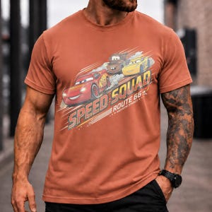 Op de afbeelding: Roestkleurig T-shirt met een afbeelding van de Cars-filmfiguren: Bliksem McQueen, Takel en een gele auto. De tekst "SPEED SQUAD ROUTE 66 RADIATOR SPRINGS" staat op de voorkant. Het shirt heeft korte mouwen.