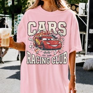 May include: Pink t-shirt featuring a graphic of a red race car with the text "CARS RACING CLUB EST. 2006". The design includes a distressed, vintage aesthetic. The shirt is a casual, oversized fit.