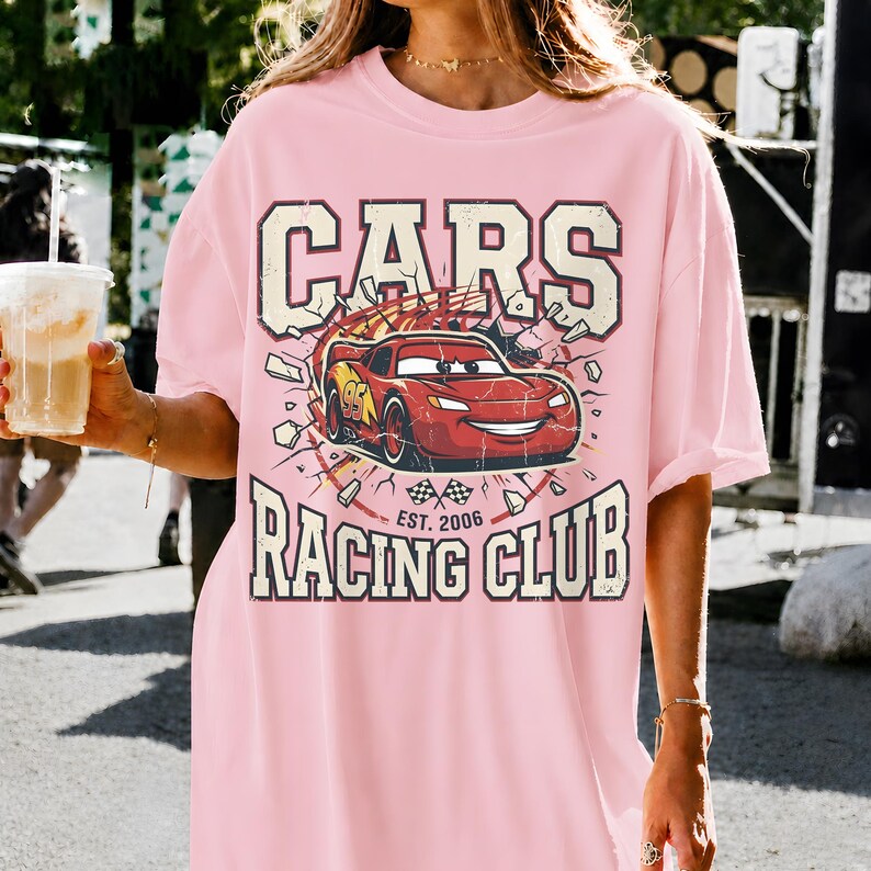 May include: Pink t-shirt featuring a graphic of a red race car with the text "CARS RACING CLUB EST. 2006". The design includes a distressed, vintage aesthetic. The shirt is a casual, oversized fit.