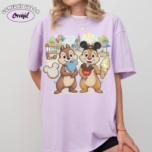 May include: A light purple Comfort Colors Orchid t-shirt featuring a cartoon illustration of Chip and Dale holding treats in front of a castle and food cart. One chipmunk holds a blue ice cream, the other a Mickey Mouse ice cream.