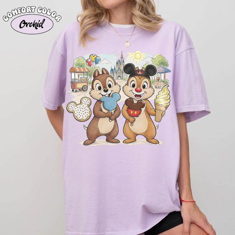 May include: A light purple Comfort Colors Orchid t-shirt featuring a cartoon illustration of Chip and Dale holding treats in front of a castle and food cart. One chipmunk holds a blue ice cream, the other a Mickey Mouse ice cream.