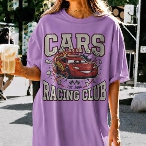 May include: A light purple t-shirt featuring a graphic of the animated character Lightning McQueen from the movie "Cars." The design includes the words "CARS" and "RACING CLUB" with "EST. 2006" below the car image.