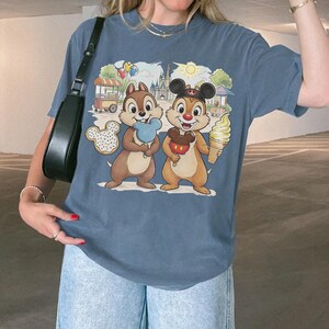 May include: Blue t-shirt featuring Chip and Dale holding treats in front of a theme park backdrop. Chip holds a blue cotton candy, and Dale holds an ice cream cone. A Mickey Mouse-shaped donut is also visible.