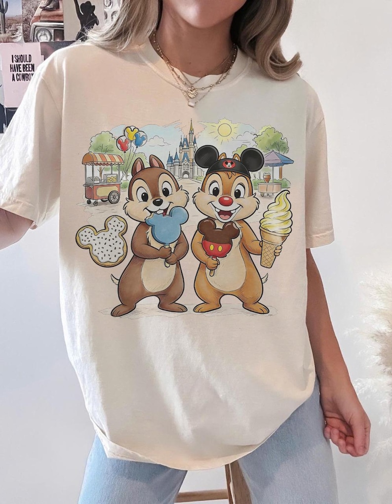 May include: Cream-colored t-shirt featuring a colorful cartoon scene with Chip and Dale holding treats in front of a castle and food stands. The shirt has a relaxed fit and short sleeves, perfect for casual wear.