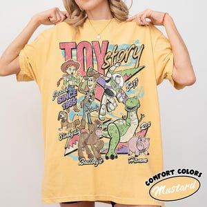May include: A mustard yellow t-shirt featuring a graphic print of Toy Story characters including Jessie, Woody, Buzz Lightyear, Slinky Dog, Bullseye, Rex, and Hamm. The text reads Toy Story, Jessie Since 1995, Woody, Buzz, Slinky Dog, Bullseye, and Hamm.