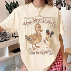 Może przedstawiać: Kremowa koszulka z nadrukiem w stylu vintage przedstawiającym dwie kaczki. Projekt zawiera tekst "Main Street Ducks Co." i "Here For The Snacks." Jedna kaczka nosi kapelusz w stylu Myszki Miki. Koszulka ma luźny krój.
