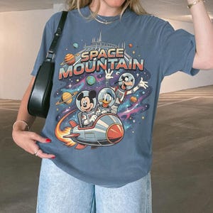 May include: Blue t-shirt featuring a colorful graphic of Mickey Mouse, Donald Duck, and Goofy in space suits on a rocket ship, with the words "SPACE MOUNTAIN" above them. The design includes planets and a starry background.