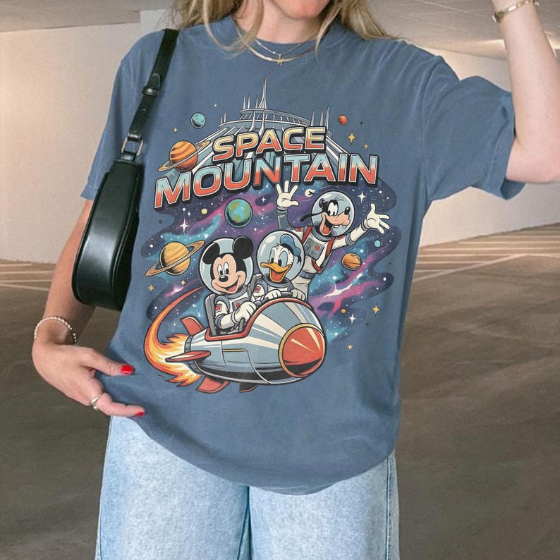 May include: Blue t-shirt featuring a colorful graphic of Mickey Mouse, Donald Duck, and Goofy in space suits on a rocket ship, with the words "SPACE MOUNTAIN" above them. The design includes planets and a starry background.
