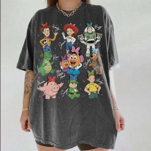 May include: Dark gray t-shirt featuring a colorful graphic of characters from the animated film Toy Story. The design includes Woody, Jessie, Buzz Lightyear, Rex, Mr. Potato Head, Slinky, Hamm, Sarge, and Andy. The names of the characters are written below them.