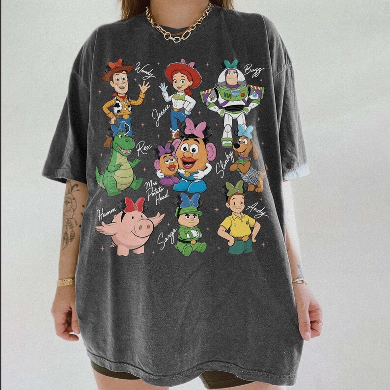 May include: Dark gray t-shirt featuring a colorful graphic of characters from the animated film Toy Story. The design includes Woody, Jessie, Buzz Lightyear, Rex, Mr. Potato Head, Slinky, Hamm, Sarge, and Andy. The names of the characters are written below them.