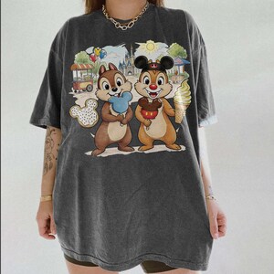 May include: Dark gray t-shirt featuring a colorful cartoon graphic of Chip and Dale holding treats in front of a theme park backdrop. The graphic includes a Mickey Mouse-shaped cookie, ice cream, and a castle.