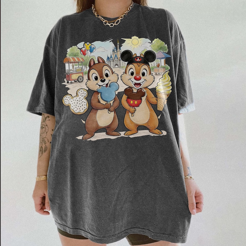 May include: Dark gray t-shirt featuring a colorful cartoon graphic of Chip and Dale holding treats in front of a theme park backdrop. The graphic includes a Mickey Mouse-shaped cookie, ice cream, and a castle.