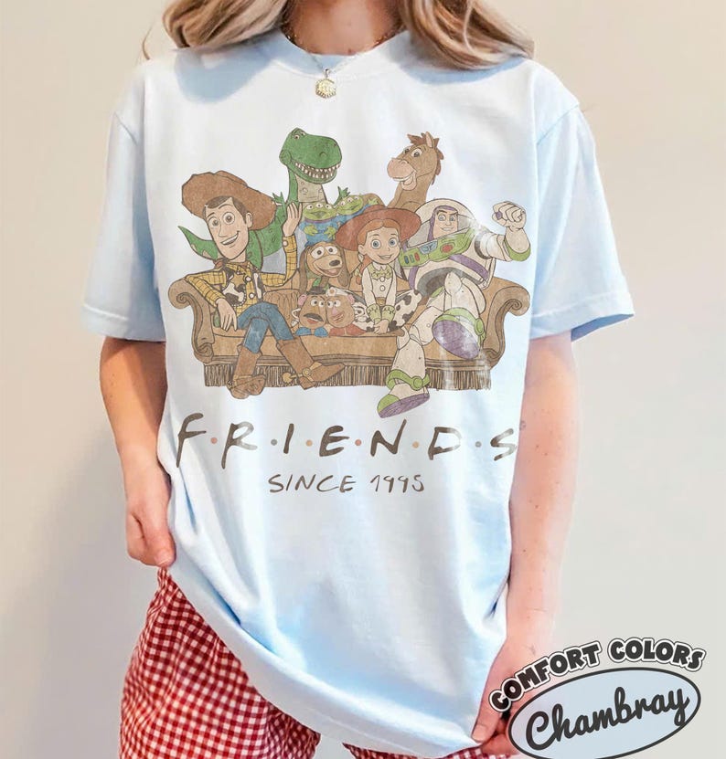 May include: A light blue t-shirt featuring a graphic of Toy Story characters sitting on a couch with the text "FRIENDS SINCE 1995" printed below. The characters include Woody, Rex, Bullseye, Slinky Dog, Mr. Potato Head, Jessie, and Buzz Lightyear.
