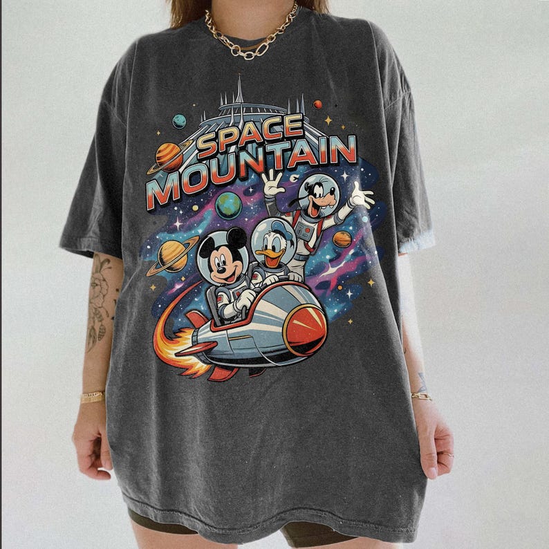 May include: Dark gray t-shirt featuring a colorful graphic of Mickey Mouse, Donald Duck, and Goofy in space suits riding a rocket ship. The words "SPACE MOUNTAIN" are printed above the characters, with planets and stars in the background.