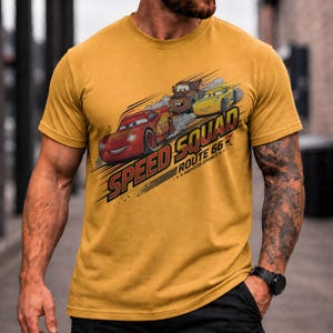 Op de afbeelding: Mosterdgeel T-shirt met een afbeelding van drie cartoon raceauto's en de tekst "Speed Squad Route 66 Radiator Springs". Het ontwerp bevat een rode auto, een bruine auto en een gele auto.