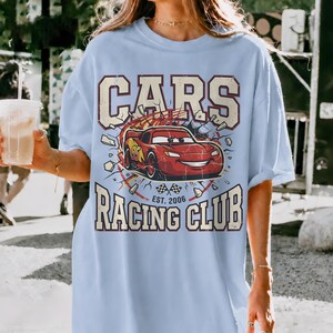 May include: Light blue t-shirt featuring a graphic of the Cars movie's Lightning McQueen. The design includes the text "CARS" above the car and "RACING CLUB EST. 2006" below. The shirt has short sleeves and a relaxed fit.