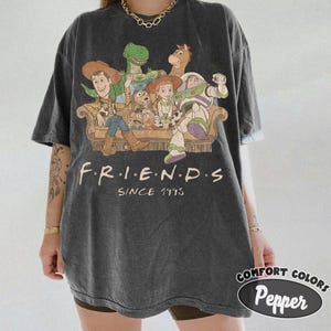 May include: A charcoal gray short-sleeve t-shirt featuring a graphic print of various animated toy characters sitting on a couch. Below the characters, the text reads F.R.I.E.N.D.S SINCE 1995 in a cream-colored font. The shirt has a crew neckline.
