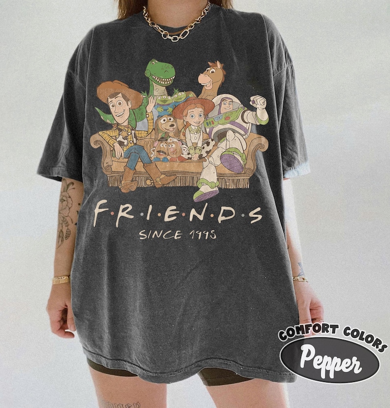 May include: A charcoal gray short-sleeve t-shirt featuring a graphic print of various animated toy characters sitting on a couch. Below the characters, the text reads F.R.I.E.N.D.S SINCE 1995 in a cream-colored font. The shirt has a crew neckline.