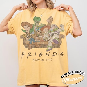 May include: A mustard yellow short-sleeve t-shirt featuring a graphic print of Toy Story characters sitting on a couch. The text "F.R.I.E.N.D.S" and "SINCE 1995" is printed below the illustration in a dark brown, stylized font.