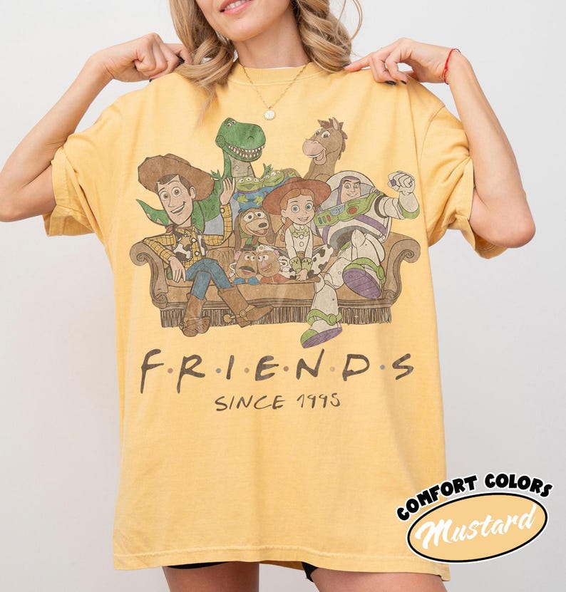 May include: A mustard yellow short-sleeve t-shirt featuring a graphic print of Toy Story characters sitting on a couch. The text "F.R.I.E.N.D.S" and "SINCE 1995" is printed below the illustration in a dark brown, stylized font.