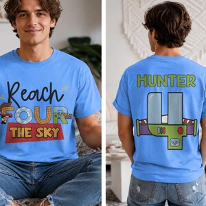 以下が含まれることがあります： 「Reach FOUR THE SKY」の文字と漫画のグラフィックが入った水色のTシャツ。Tシャツの背面には、漫画のグラフィックの上に「HUNTER」という名前が描かれています。Tシャツは人が着ています。